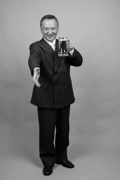 Зйомки дорослого азіатського бізнесмена, який тримає келих пива на сірому фоні в чорно-білому - Фото, зображення