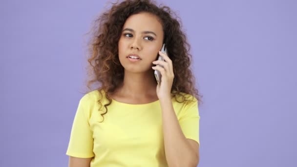 African young woman in yellow t-shirt having an annoying call while looking at the camera over purple background isolated - Кадри, відео