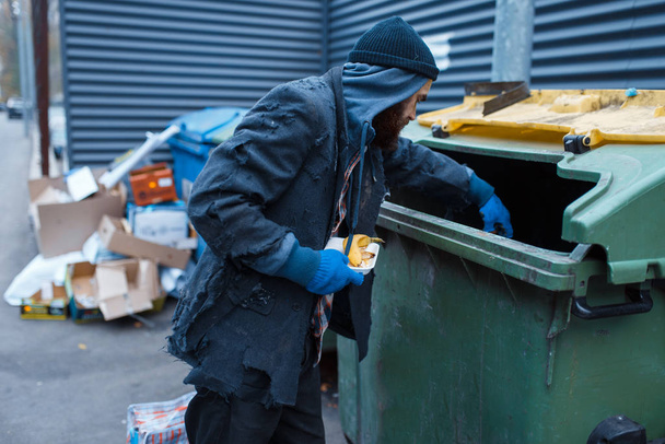 Мужчина бородатый нищий ищет еду в мусорном баке на городской улице. Бедность - социальная проблема, бездомность и одиночество, алкоголизм и пьянство
 - Фото, изображение