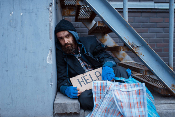 Bearded bum with help sign on city street. Poverty is a social problem, homelessness and loneliness, alcoholism and  drunk addiction, urban lonely - Foto, Imagem