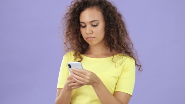 Calm pretty african young woman in yellow t-shirt writes something on a smartphone over purple background isolated - Filmagem, Vídeo
