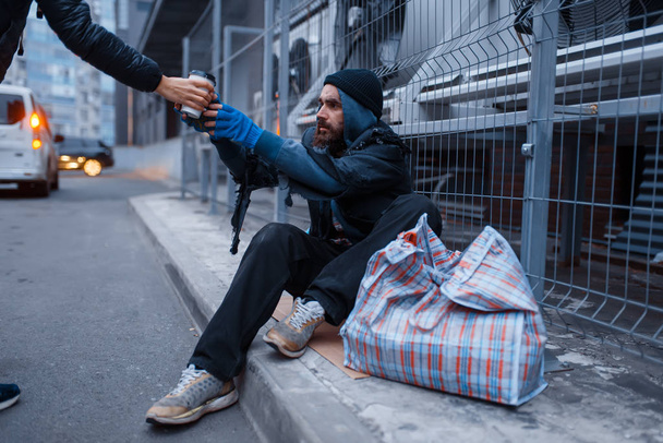 Male person gives cup of hot coffee to bearded dirty homeless on city street. Poverty is a social problem, homelessness and loneliness, alcoholism and  drunk addiction, urban lonely - Photo, image