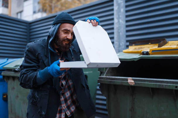 Bearded dirty beggar found pizza in trashcan on city street. Poverty is a social problem, homelessness and loneliness, alcoholism and  drunk addiction, urban lonely - Photo, Image