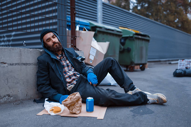 Bearded dirty beggar with food sitting at the trashcan on city street. Poverty is a social problem, homelessness and loneliness, alcoholism and  drunk addiction, urban lonely - Photo, Image