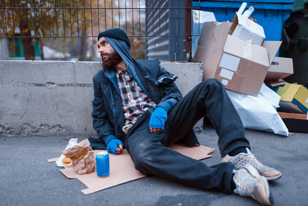 Bearded dirty beggar with food sitting at the trashcan on city street. Poverty is a social problem, homelessness and loneliness, alcoholism and drunk addiction - Photo, Image