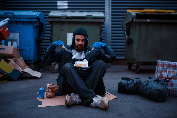 Vuile bedelaar met mes en vork eet in de vuilnisbak op straat. Armoede is een sociaal probleem, dakloosheid en eenzaamheid, alcoholisme en alcoholverslaving, stedelijke eenzaamheid - Foto, afbeelding