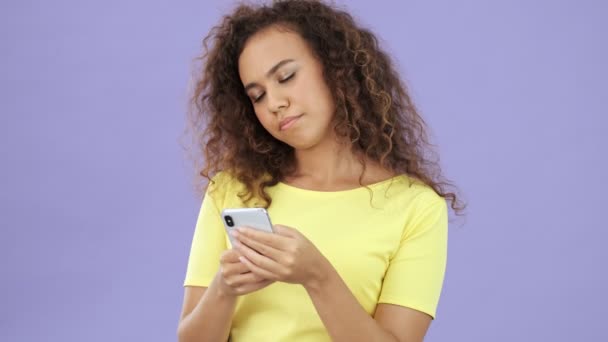 Beautiful african young woman in yellow t-shirt becoming displeased while chatting on smartphone over purple background isolated - Кадри, відео