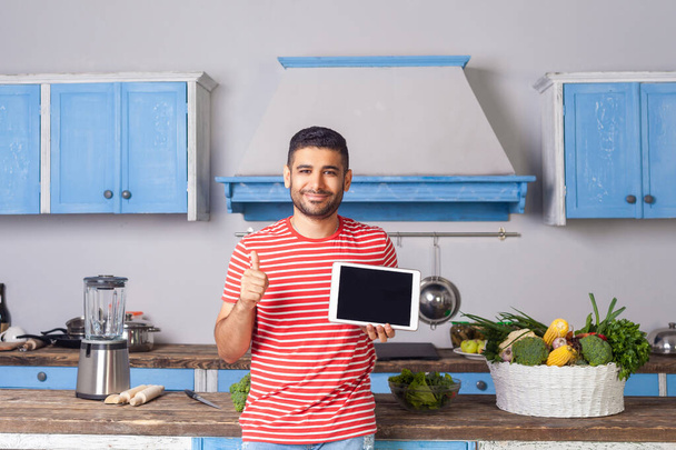 Happy satisfied man standing in kitchen, holding tablet and show - Valokuva, kuva