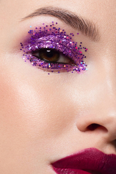 Close-up of the beauty of half a woman's face with creative fashionable make-up of sparkles. Black eyeliner and long eyelashes decorate hazel eyes and lips to match. Well-groomed skin after spa - Foto, afbeelding