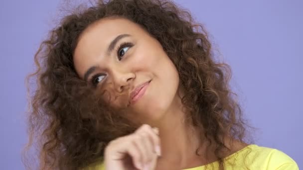 Close up view of cute flirty african young woman in yellow t-shirt smiling and playing with her hair while looking at the camera over purple background isolated - Imágenes, Vídeo