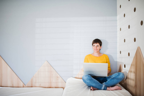 Young woman with laptop sitting on bed in bedroom indoors at home. - Fotografie, Obrázek