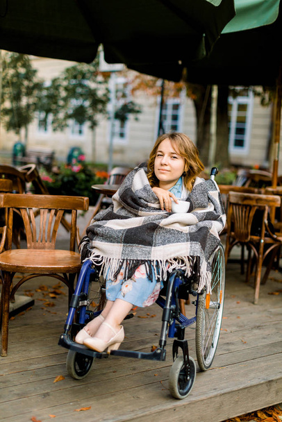 Portrait of a young beautiful woman on a wheelchair, covered with checkered plaid, drinking coffee while sitting in cafe outdoors. - Foto, afbeelding
