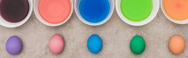 top view of painted chicken eggs near watercolor paints in bowls on grey concrete surface, panoramic shot - Photo, Image