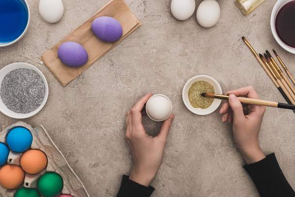 cropped view of woman decorating chicken eggs with golden glitter on grey concrete surface - Foto, Bild