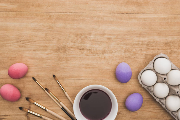top view of watercolor purple paint in bowl near chicken eggs and paintbrushes on wooden table - 写真・画像