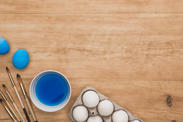 vista superior de acuarela pintura azul en tazón cerca de huevos de pollo y pinceles en mesa de madera
 - Foto, imagen