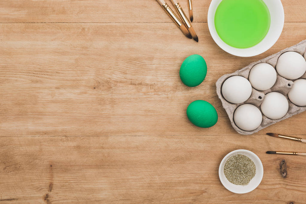 Paskalya yumurtaları ve ahşap masadaki boya fırçalarının yanındaki kaselerde yeşil suluboya ve parıltı manzarası. - Fotoğraf, Görsel