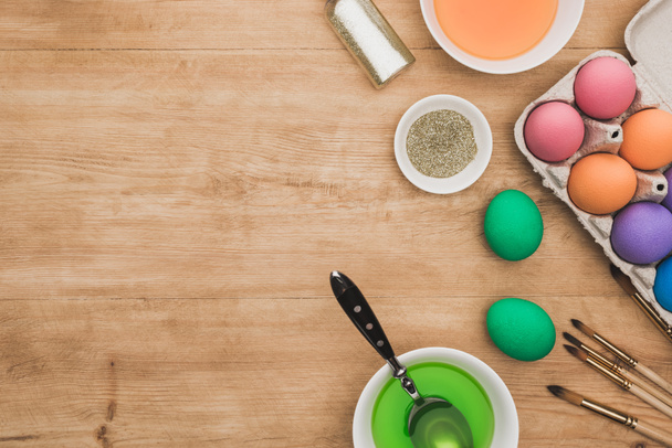 vrchní pohled na akvarelové barvy a třpytky v miskách v blízkosti velikonočních vajec a štětců na dřevěném stole - Fotografie, Obrázek