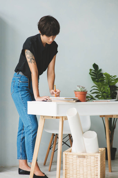 Young business woman standing near the desk and taking notes in notebook. - Foto, imagen