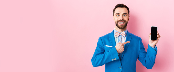 plano panorámico de hombre de negocios barbudo feliz apuntando con el dedo al teléfono inteligente con pantalla en blanco en rosa
  - Foto, imagen