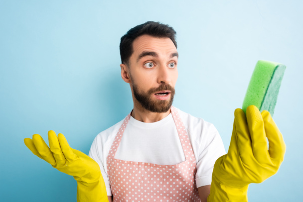 selective focus of surprised man in dotted apron looking at sponge on blue  - 写真・画像