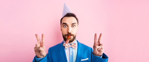 panoramic shot of businessman in party cap holding party blower in mouth and showing peace sign isolated on pink  - Foto, Imagen