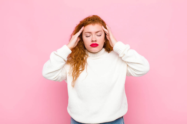 young red head woman looking concentrated, thoughtful and inspired, brainstorming and imagining with hands on forehead against pink wall - 写真・画像