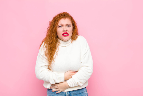 young red head woman feeling anxious, ill, sick and unhappy, suffering a painful stomach ache or flu against pink wall - Fotografie, Obrázek