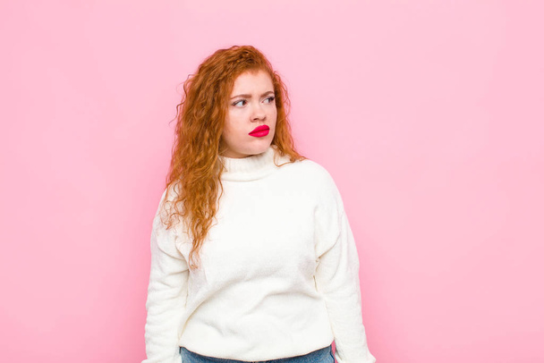 young red head woman feeling sad, upset or angry and looking to the side with a negative attitude, frowning in disagreement against pink wall - 写真・画像