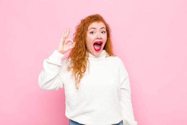young red head woman feeling successful and satisfied, smiling with mouth wide open, making okay sign with hand against pink wall - 写真・画像