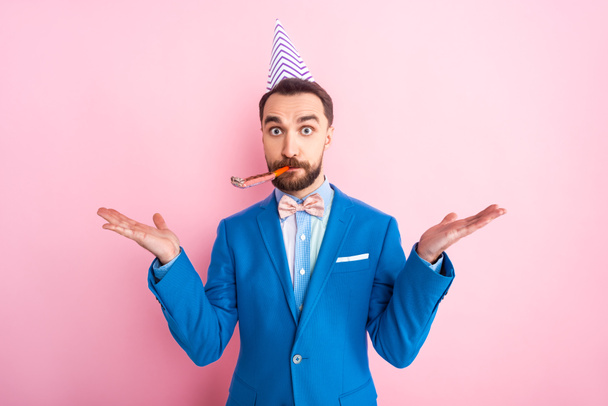 confused businessman in party cap showing shrug gesture isolated on pink  - Foto, Bild
