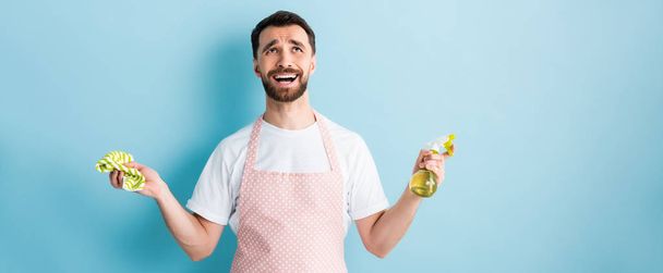 panoramic shot of emotional man in apron holding rag and spray bottle on blue  - 写真・画像