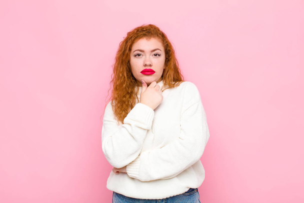 young red head woman looking serious, confused, uncertain and thoughtful, doubting among options or choices against pink wall - Фото, зображення