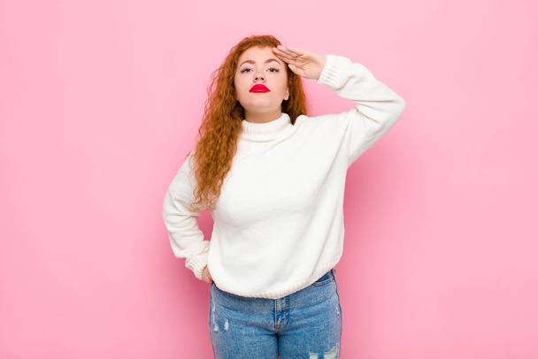 young red head woman greeting the camera with a military salute in an act of honor and patriotism, showing respect against pink wall - Foto, imagen