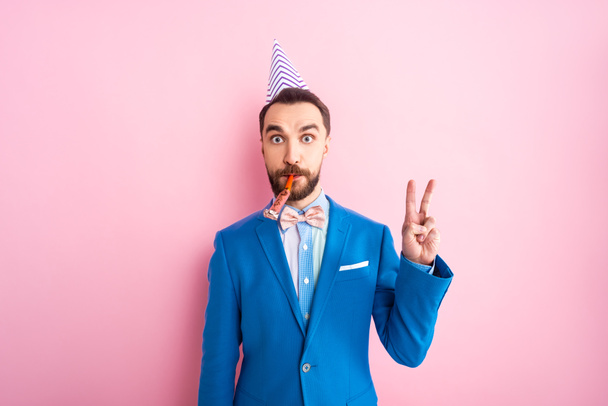 businessman in party cap blowing party blower and showing peace sign on pink  - Foto, Imagem