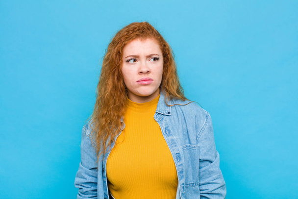 young red head woman feeling sad, upset or angry and looking to the side with a negative attitude, frowning in disagreement against blue wall - Foto, afbeelding
