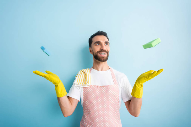 cheerful bearded man in apron throwing in air sponges on blue  - Foto, Imagem