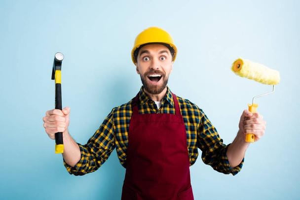 excited workman holding hammer and paint roller on blue  - Zdjęcie, obraz