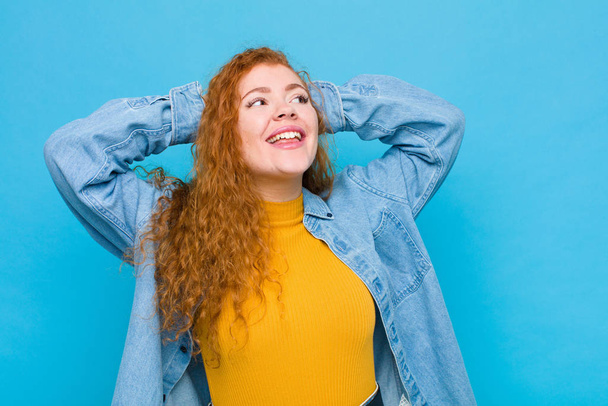 young red head woman smiling and feeling relaxed, satisfied and carefree, laughing positively and chilling against blue wall - Foto, afbeelding