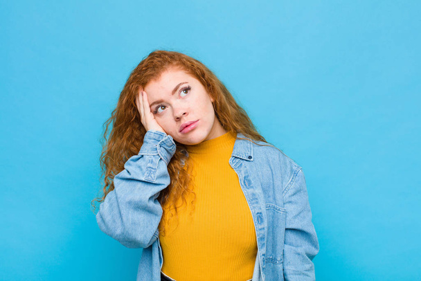 young red head woman feeling bored, frustrated and sleepy after a tiresome, dull and tedious task, holding face with hand against blue wall - Φωτογραφία, εικόνα