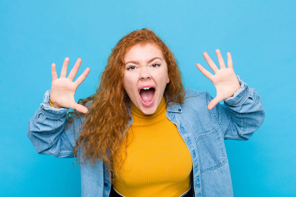 young red head woman screaming in panic or anger, shocked, terrified or furious, with hands next to head against blue wall - Фото, изображение