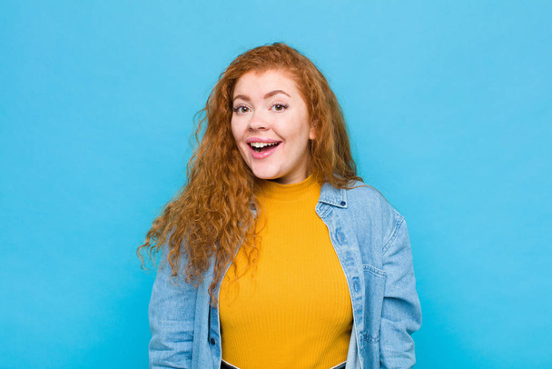 young red head woman looking happy and pleasantly surprised, excited with a fascinated and shocked expression against blue wall - Photo, image