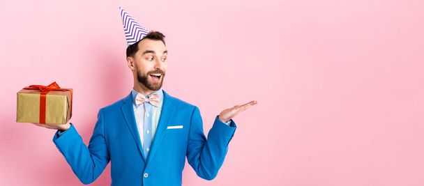 panoramic shot of happy businessman holding present and pointing with hand on pink  - Foto, imagen