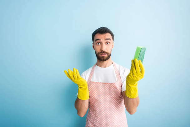 confused bearded man in rubber gloves holding sponge on blue  - Foto, Bild