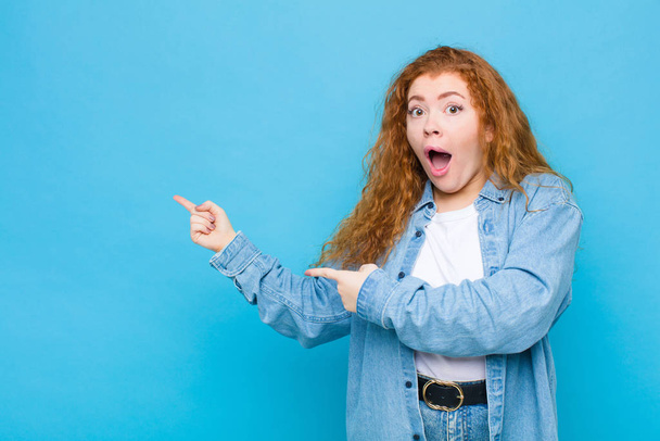 young red head woman feeling shocked and surprised, pointing to copy space on the side with amazed, open-mouthed look against blue wall - Fotó, kép