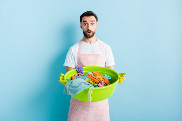 shocked bearded man holding plastic wash bowl with dirty laundry on blue  - Fotografie, Obrázek