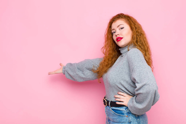 young red head woman feeling happy and cheerful, smiling and welcoming you, inviting you in with a friendly gesture against pink wall - Photo, Image