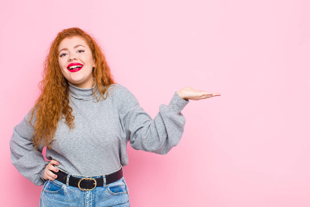 young red head woman smiling, feeling confident, successful and happy, showing concept or idea on copy space on the side against pink wall - Foto, imagen