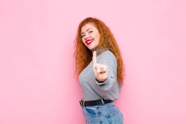 young red head woman smiling proudly and confidently making number one pose triumphantly, feeling like a leader against pink wall - Fotoğraf, Görsel
