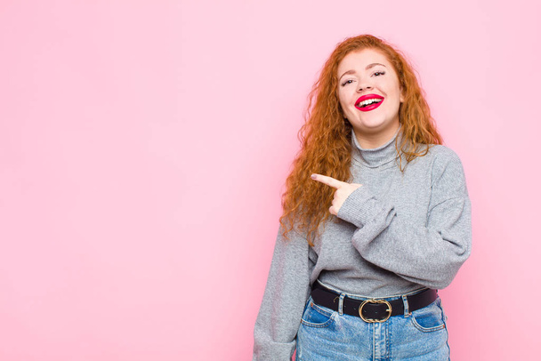 young red head woman smiling cheerfully, feeling happy and pointing to the side and upwards, showing object in copy space against pink wall - Foto, immagini